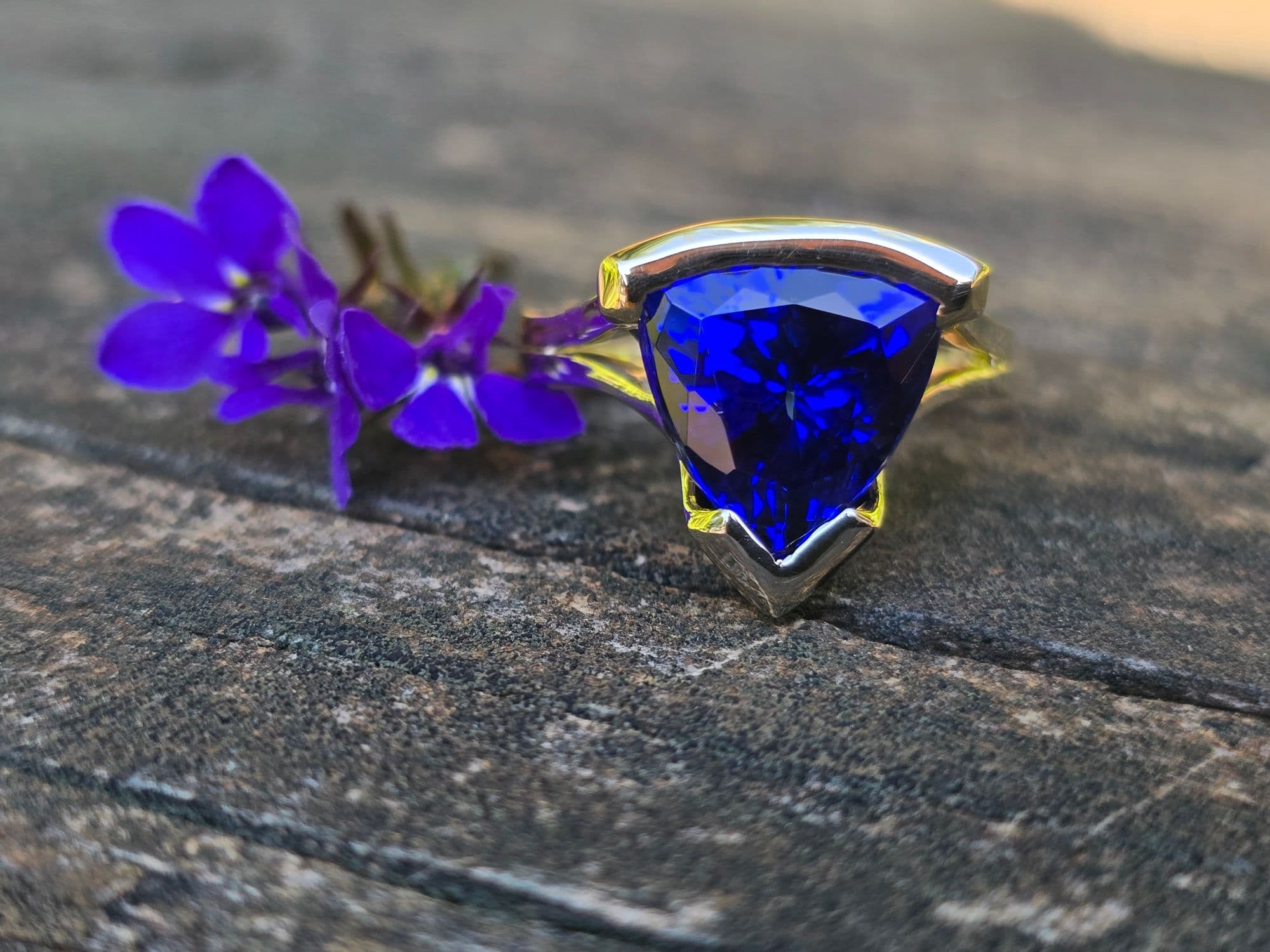Ring with a large, deep violet-blue, triangular stone held in an angular open bezel of white-toned metal on a split, yellow-toned band, beside small purple flowers