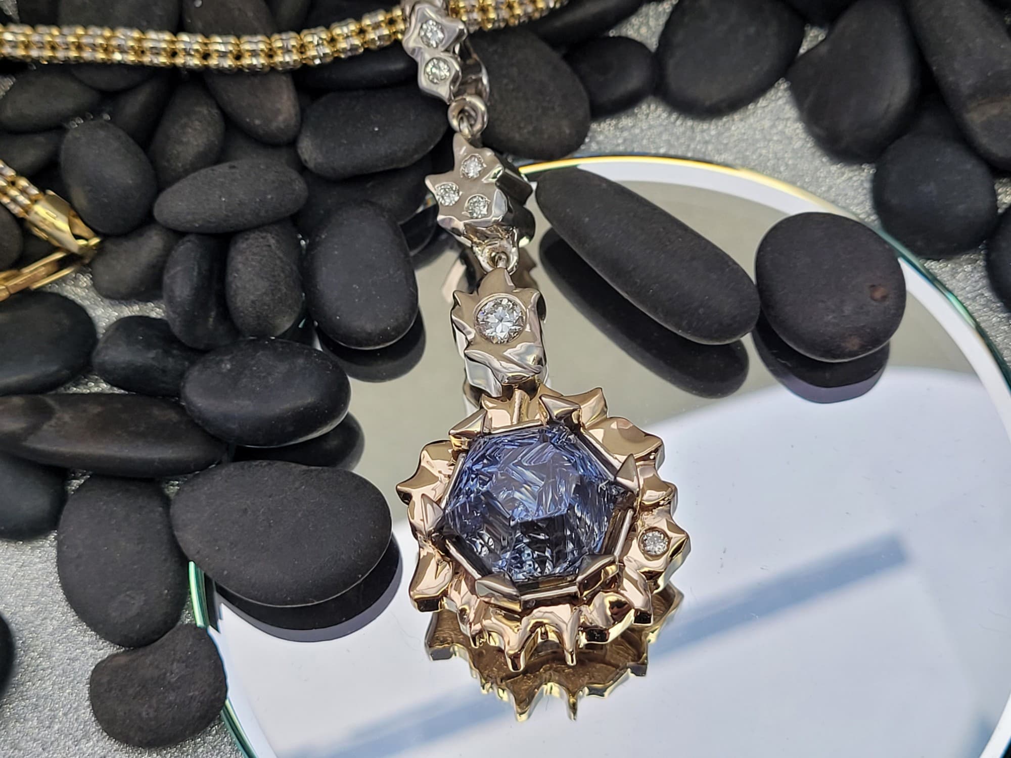 Pendant with a large blue, hexagonal stone cut with carved, frosted facets, held by tall points in a sculpted sunburst frame of rose- and yellow-toned metal, below white-toned links set with small colorless stones