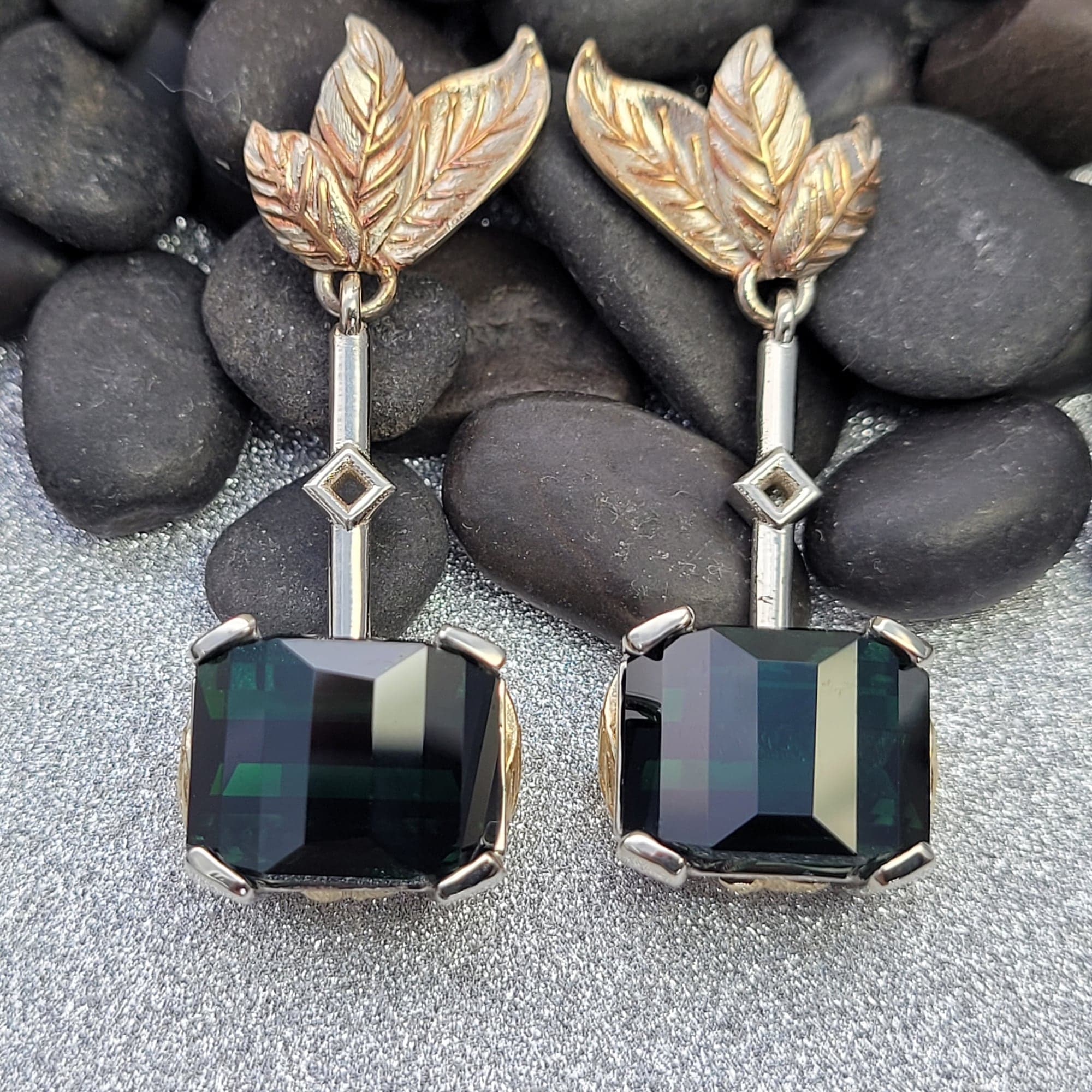 Pair of drop earrings: sculpted gold-toned leaves above a slim bar and a deep green, step-cut rectangular stone, resting on black river stones