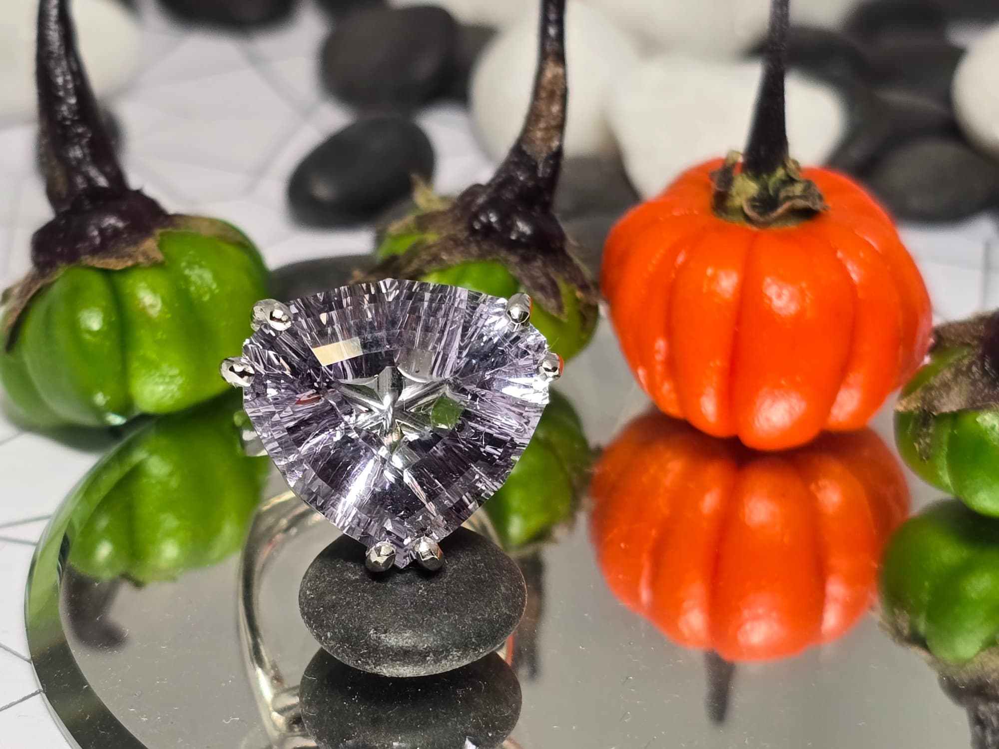 Ring with a large, pale lavender, triangular stone cut with radiating grooved facets, held by paired white-toned prongs, shown among small green and orange gourds