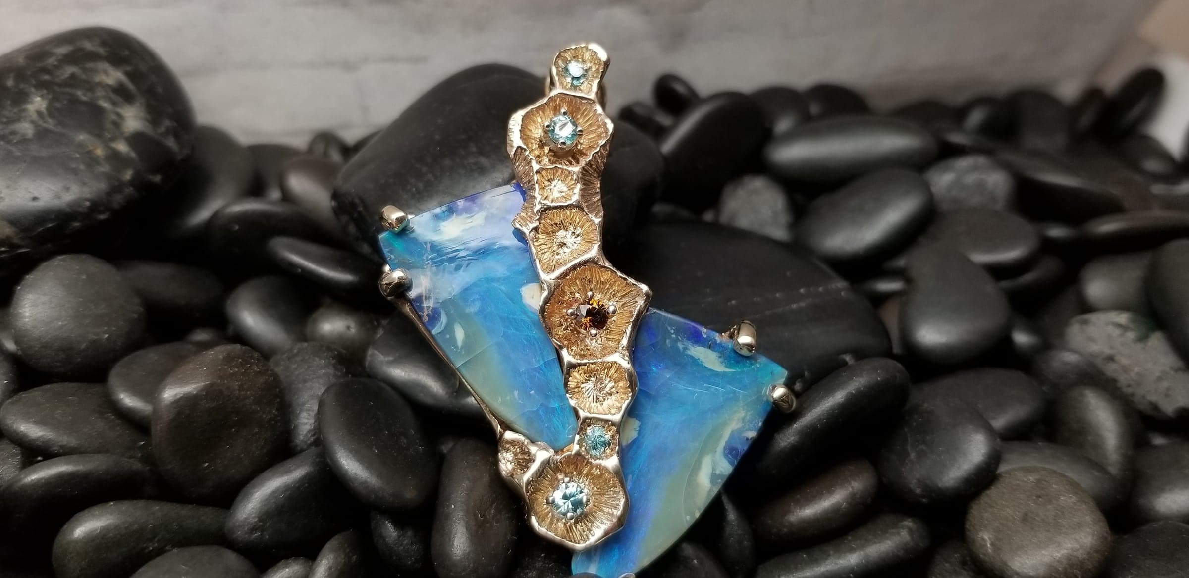 The Gilded Reef: a pendant with two triangular blue stones held by a column of sculpted, gold-toned coral forms set with small blue and amber-colored stones, resting on black river stones