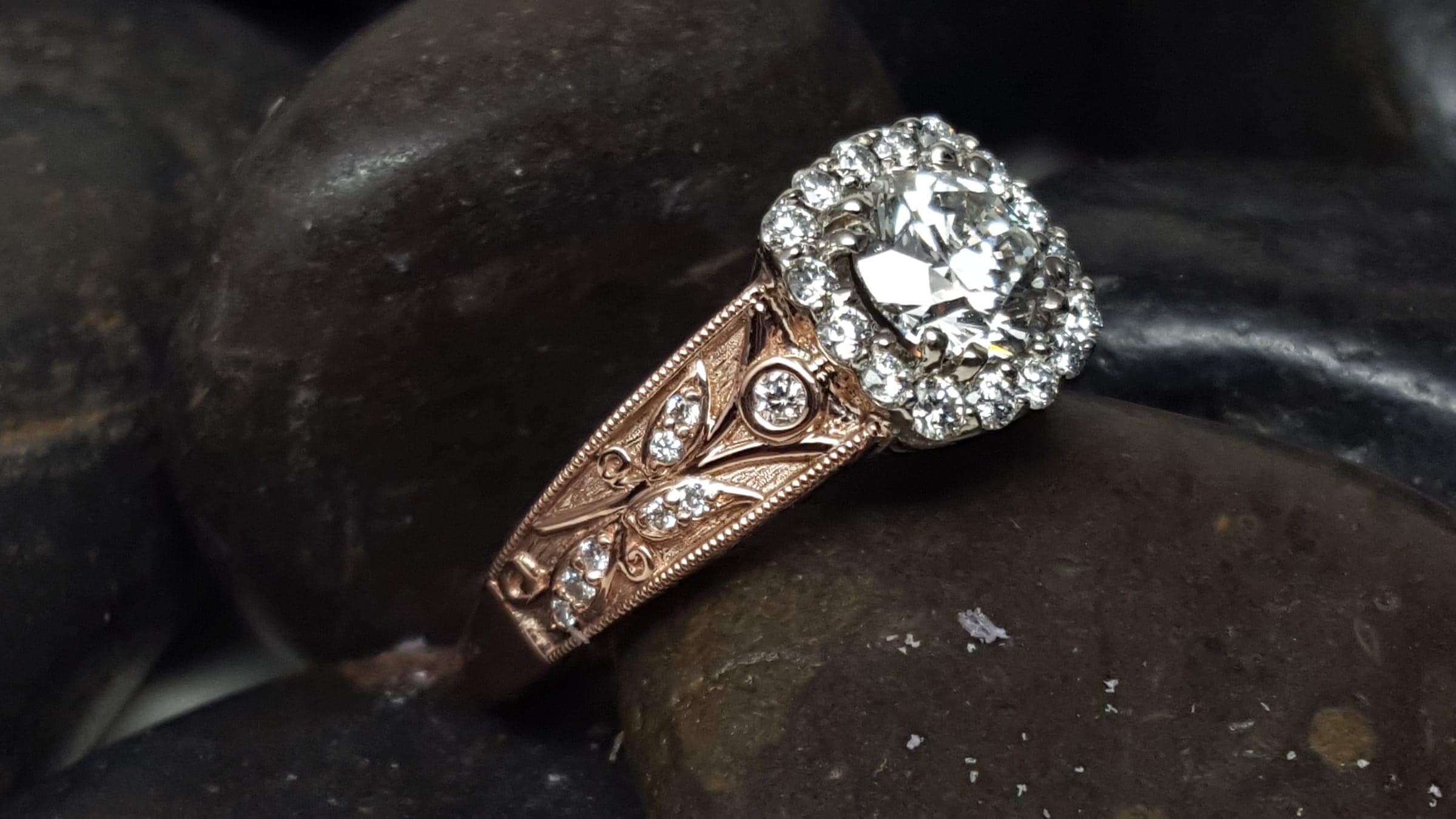 Ring with a round, colorless center stone in a halo of small colorless stones, on a rose-toned band whose shoulders are engraved with leaves and set with small colorless stones, resting on dark river stones