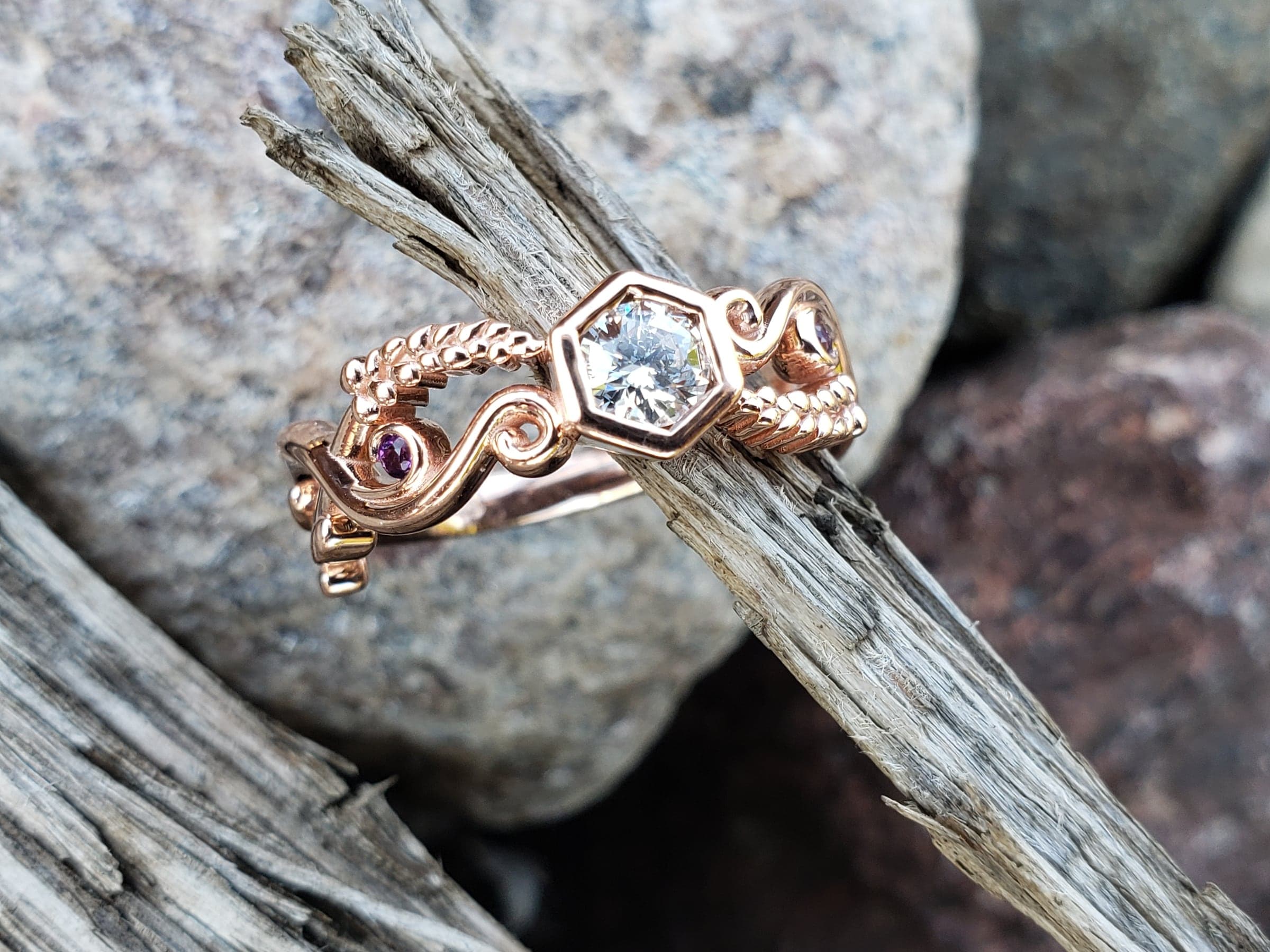 Rose-toned ring with a round, colorless stone in a hexagonal setting, rope-twist wire, open scrollwork and small purple accent stones, resting on weathered wood beside grey rocks