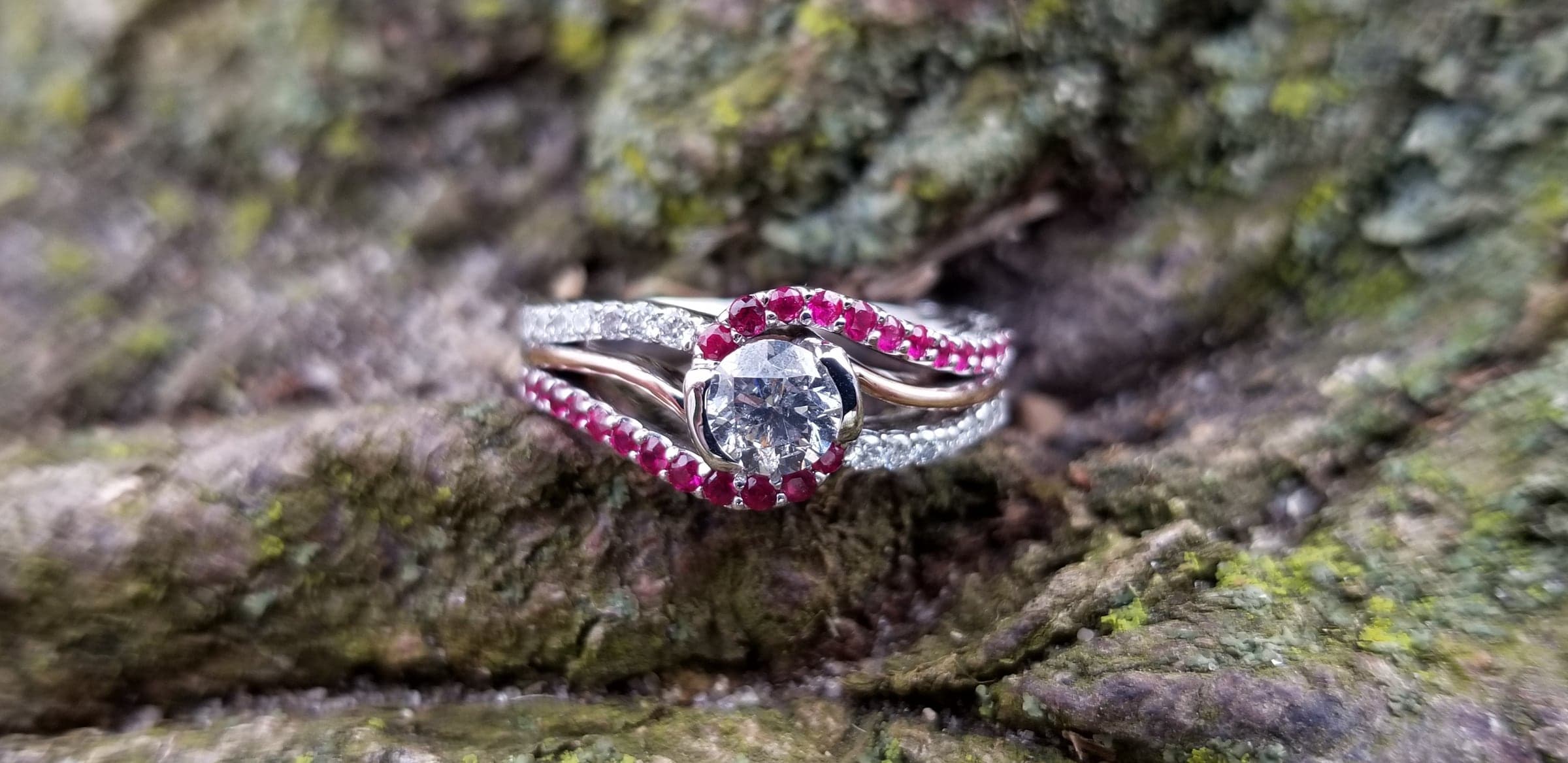 Rose gold and white gold ring: a round center stone with sweeping rows of small red and colorless stones, on moss-covered stone