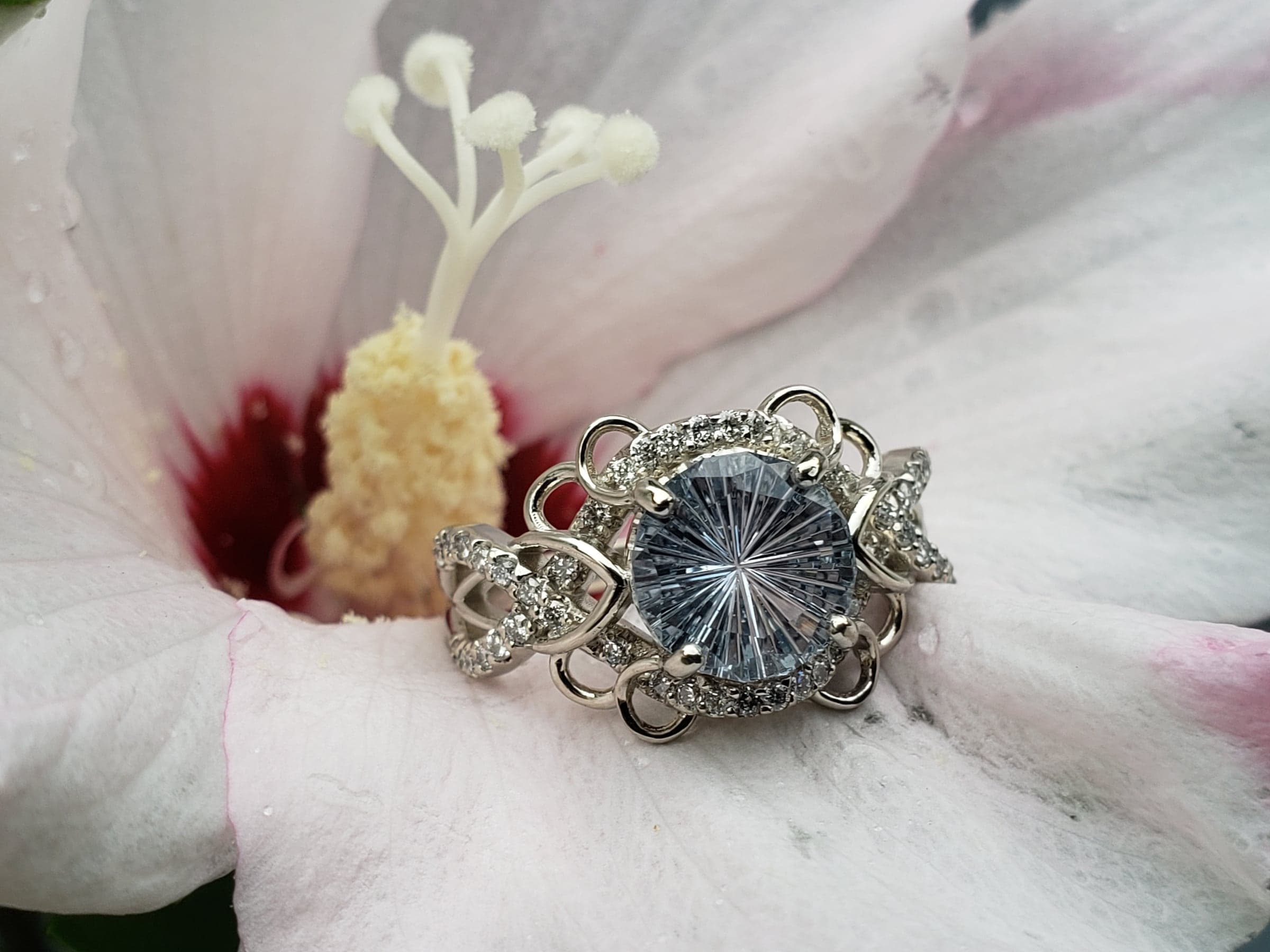 Ring with a large, round, pale blue stone cut in a starburst pattern, framed by small colorless stones in an open, looping white-toned setting, resting in a white and pink flower