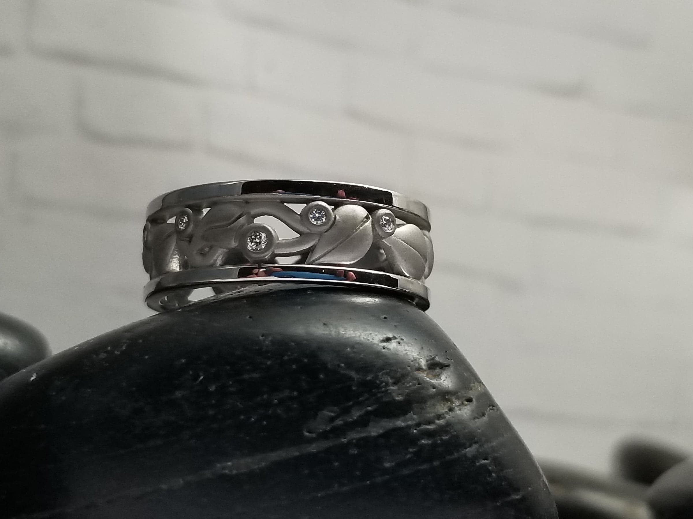 Wide white-toned band with an open pattern of sculpted leaves and curling vines between two polished rails, set with small colorless stones, standing on a dark stone