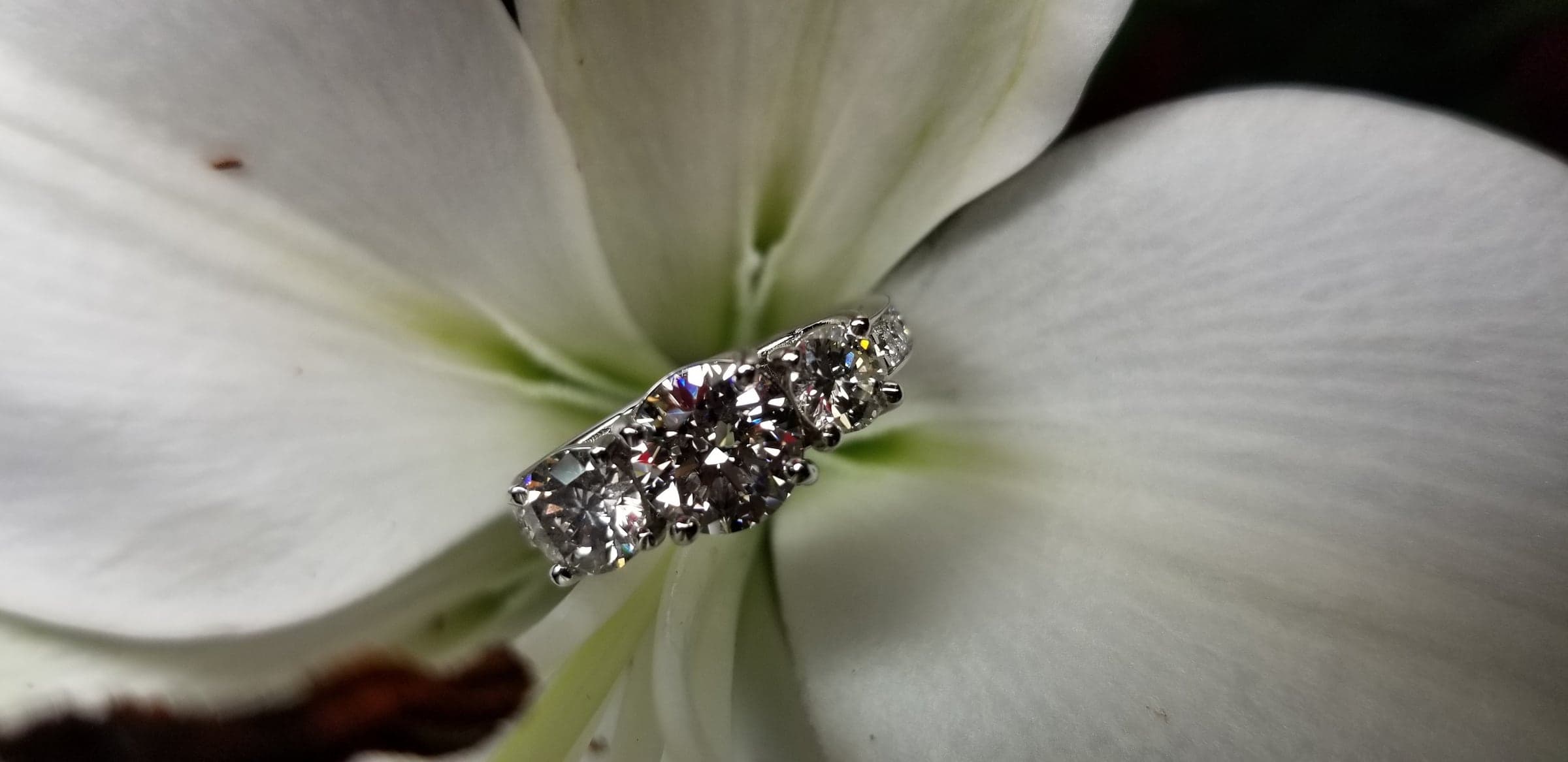 Ring with three colorless stones in a row, the center stone largest, in a white-toned setting, resting on a white flower