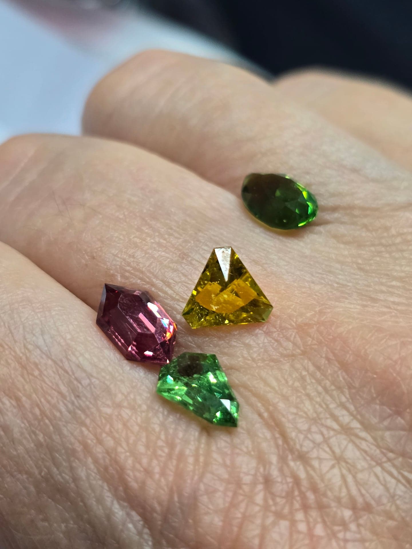 Four gemstones in pink, golden yellow and two shades of green, each in an unusual cut, resting on the back of a hand