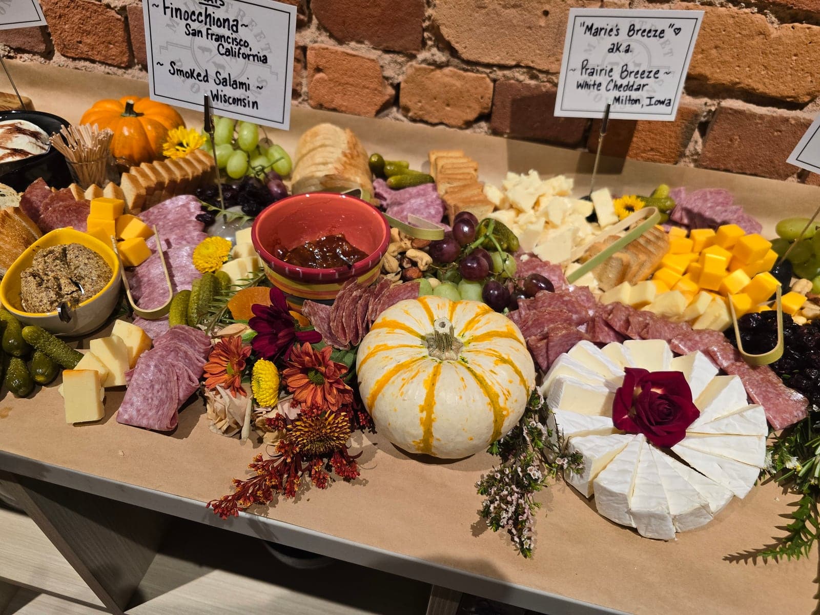 A grazing table of cheeses, salami, grapes and fresh flowers, with hand-lettered signs including “Marie’s Breeze”