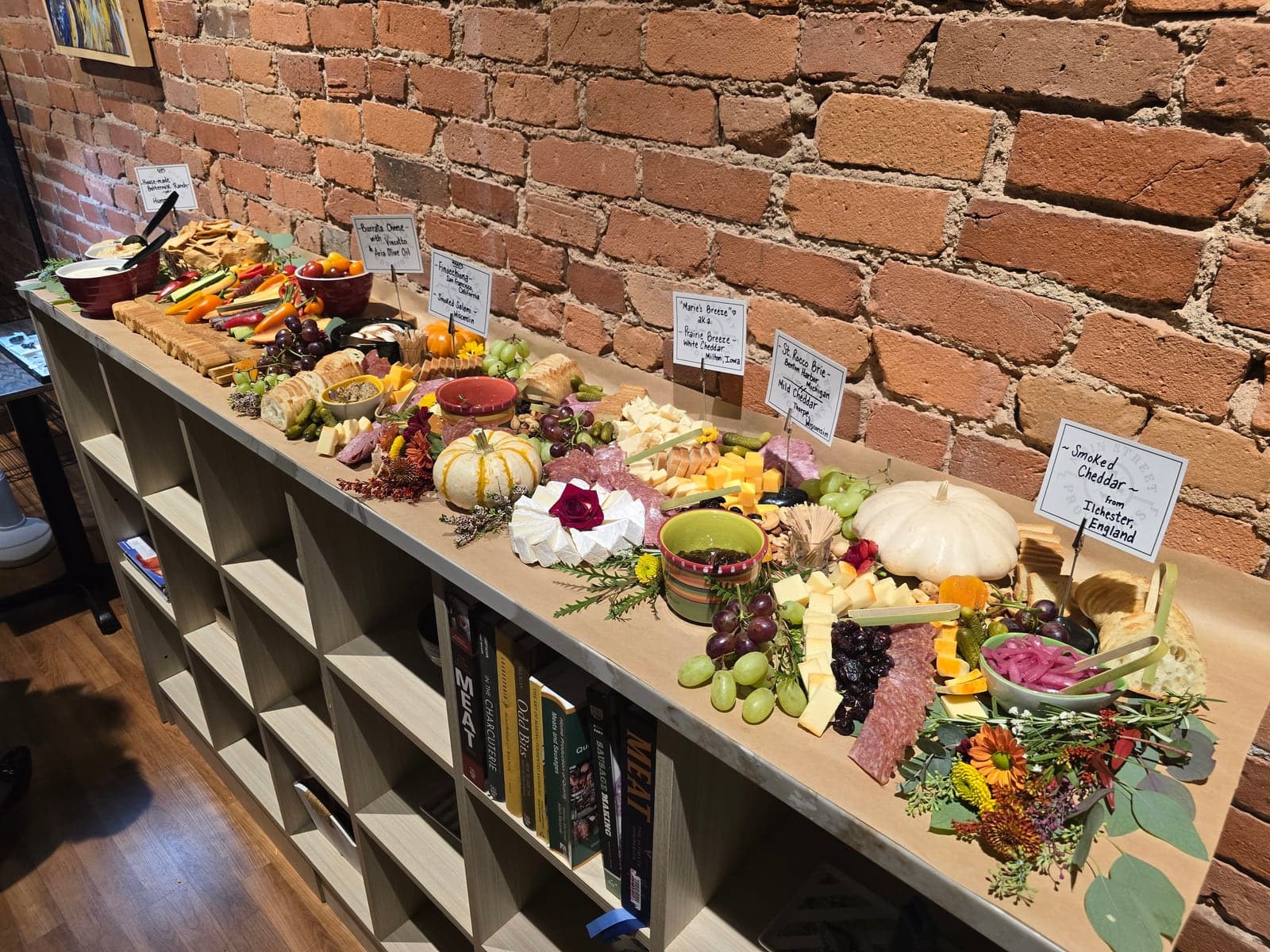 A long grazing table stretching along the brick wall, laden with cheeses, charcuterie, fruit, vegetables and flowers