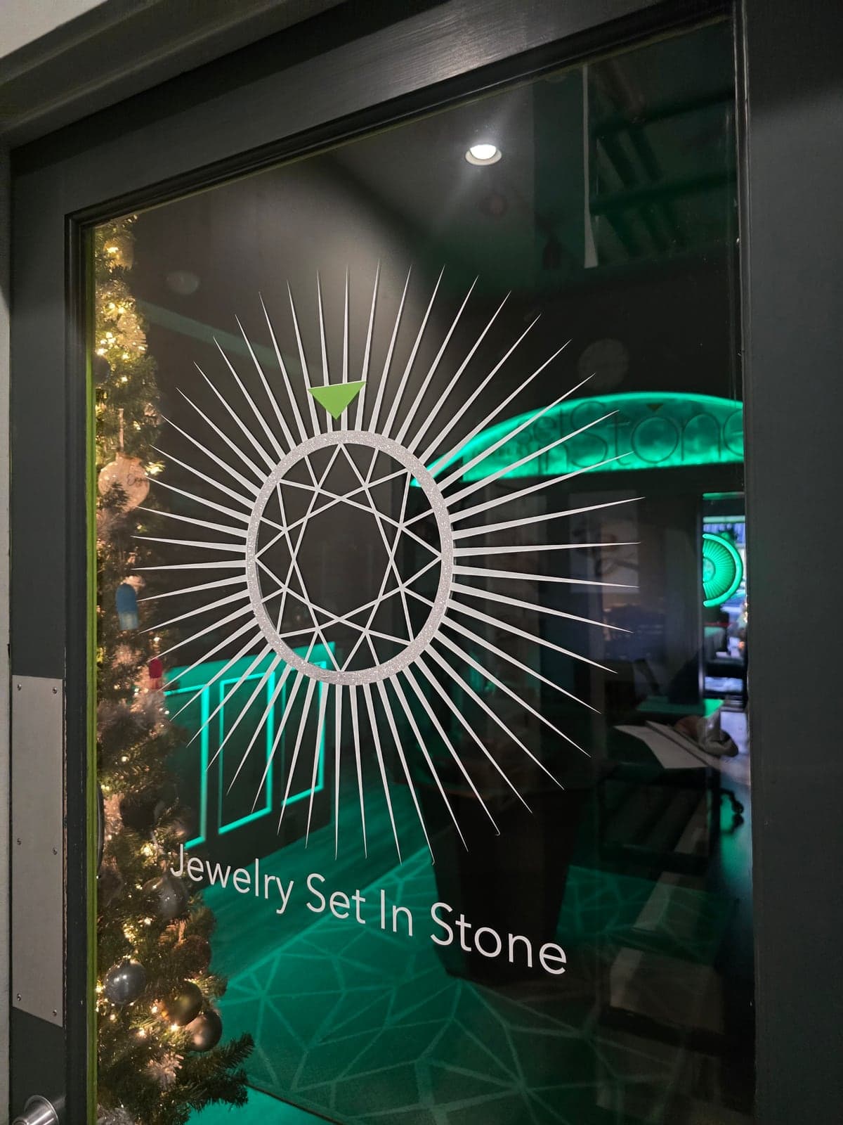 The Jewelry Set in Stone logo on the glass studio door, with a lit Christmas tree and the glowing green sign inside